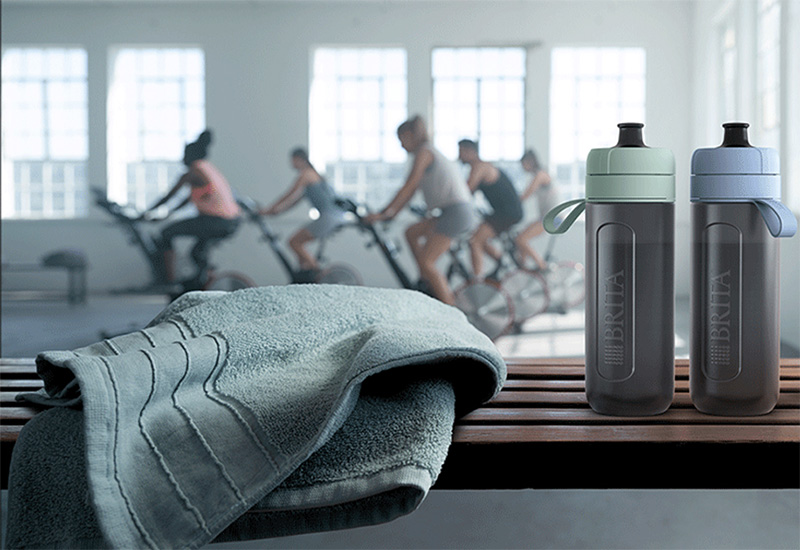 Zwei Model Actives stehen auf einer Bank im Fitnessstudio, dahinter findet ein Spinning-Kurs statt.